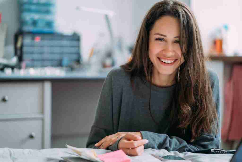 Portrait of smiling cute hipster girl staring on camera while writing college exercises in notebook