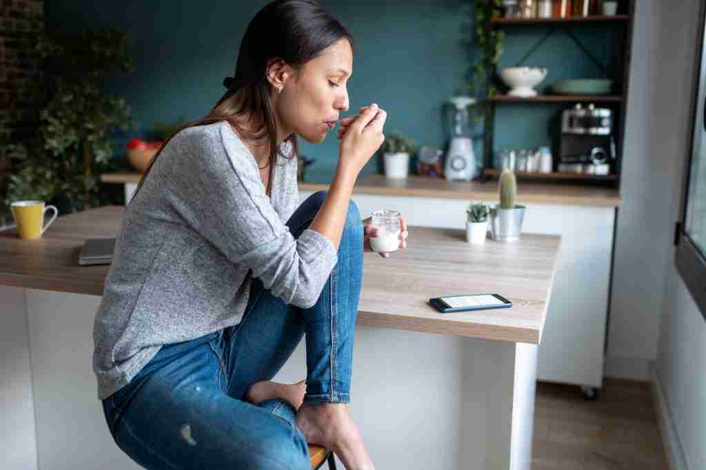 Pretty young woman eating yogurt while using her mobile phone in the kitchen at home.