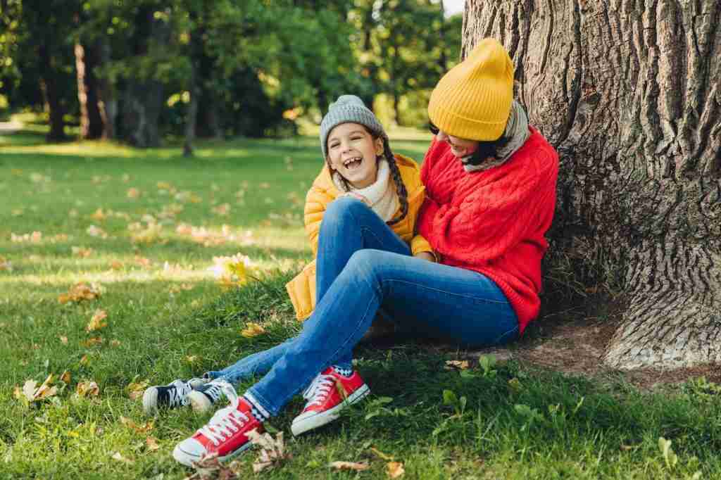 Funny little child and her mother sit together near tree, have walk in autumn park