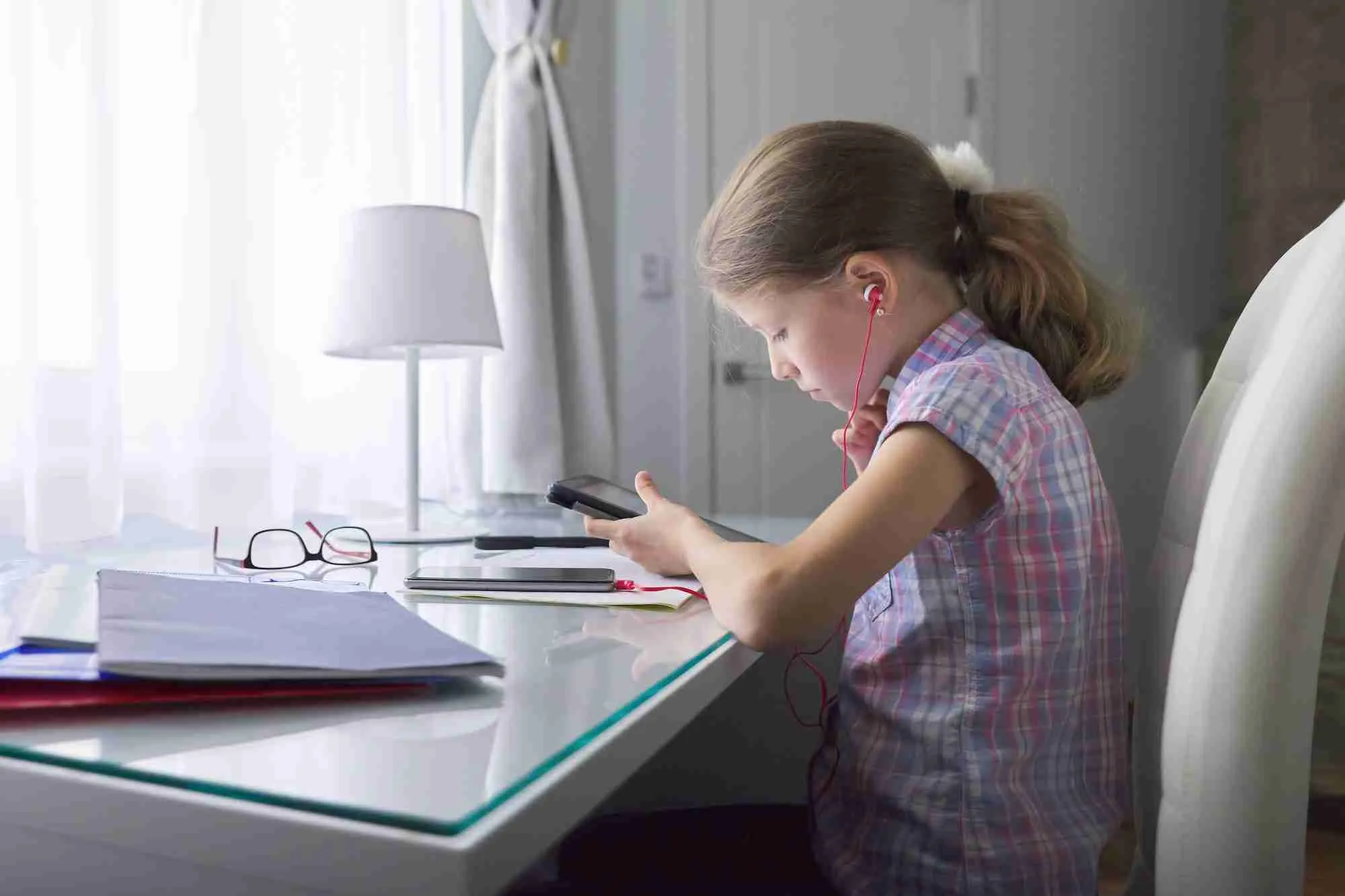 Girl child sitting at home at desk near window with school notebooks and digital tablet
