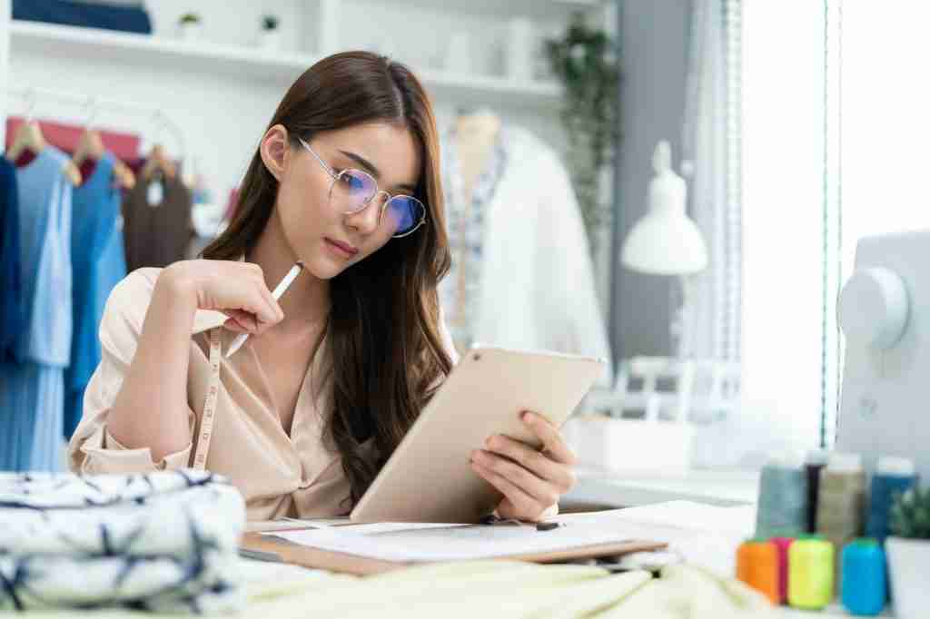 Asian tailor woman use tablet to find reference to think and design new clothes in workshop room