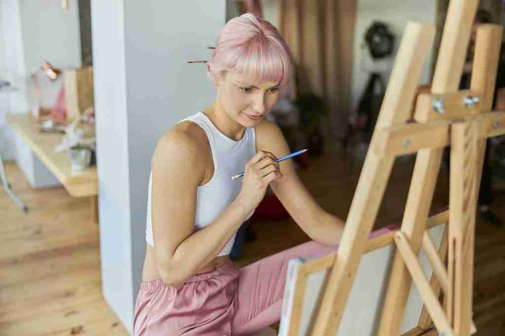 Thoughtful woman with pink hair looks at canvas holding pencil in light studio