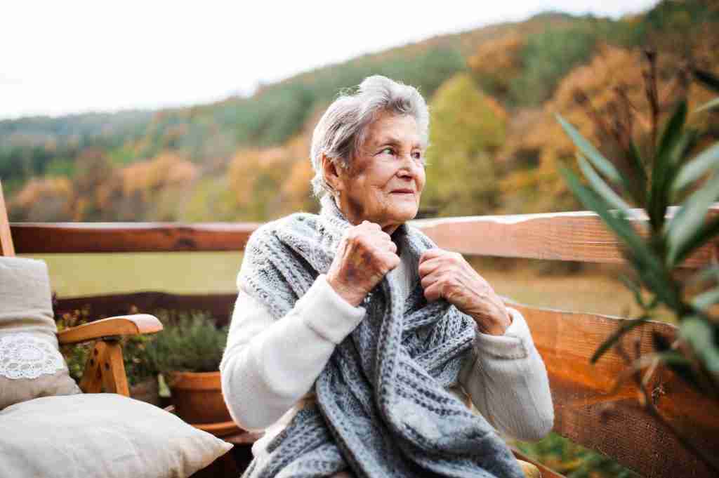 An elderly woman sitting outdoors on a terrace on a sunny day in autumn.