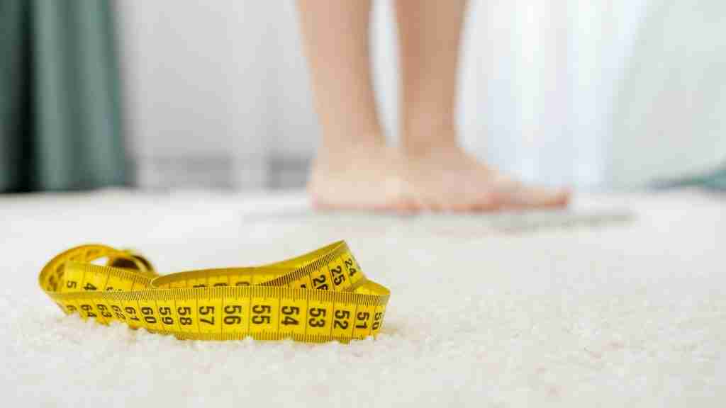 Camera focusing on measuring tape and woman weighting on scales at bedroom. Concept of dieting
