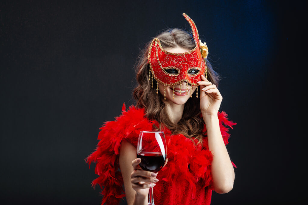Smiling young woman in a red carnival mask and boa with a raised glass of wine. - Image