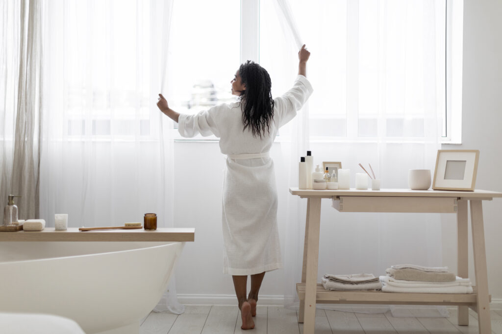 Young Black Woman Opening Curtains While Making Morning Beauty Routine In Bathroom