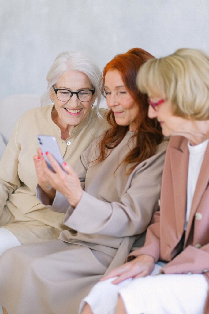Senior cheerful women in eyeglasses looking at screen of smartphone and surfing internet together
