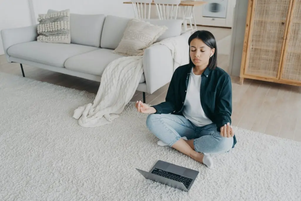 Female practices yoga at laptop on floor, listening meditation to relieve stress after work at home
