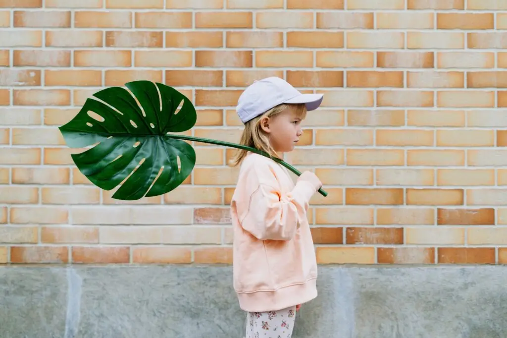 Thoughtful little girl holding monstera leaf in front of brick wall.