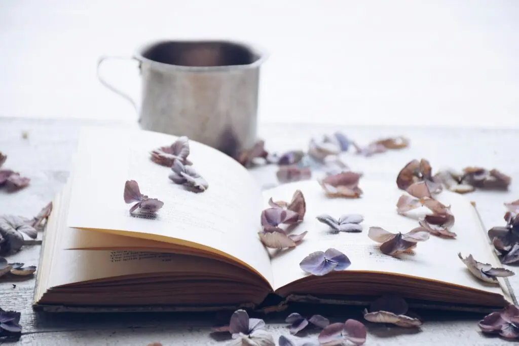An open book with scattered hydrangea flower petals and a cup of tea or coffee in the background