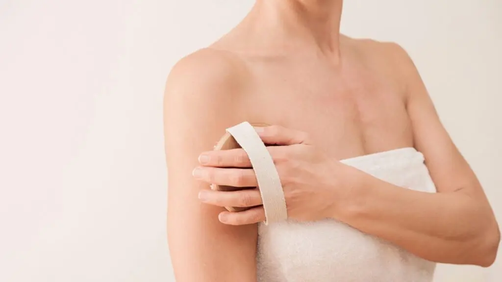 Close up of unrecognizable woman using dry brush for body exfoliation on neutral background